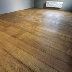 A room with a freshly installed light brown wood floor, light grey walls, and a white radiator mounted on one wall. The room is empty and well-lit, showcasing the natural wood grain of the flooring.