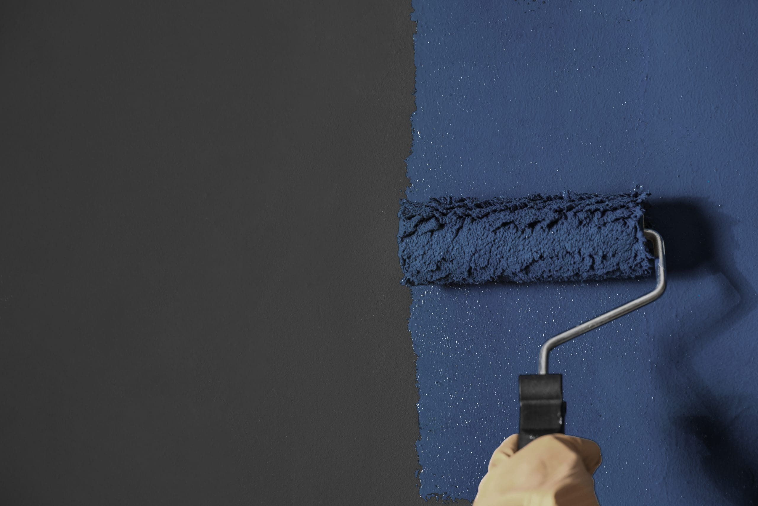 A paint roller is being used to cover a dark grey wall with a fresh coat of blue paint. The roller, held by a gloved hand, is midway through applying the new blue color.