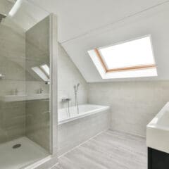 A modern bathroom featuring a glass-enclosed shower, a white bathtub with a handheld showerhead, a skylight above the tub, and a sleek white sink with a mirror. The walls and floor are tiled with light gray tiles, creating a clean and bright atmosphere.