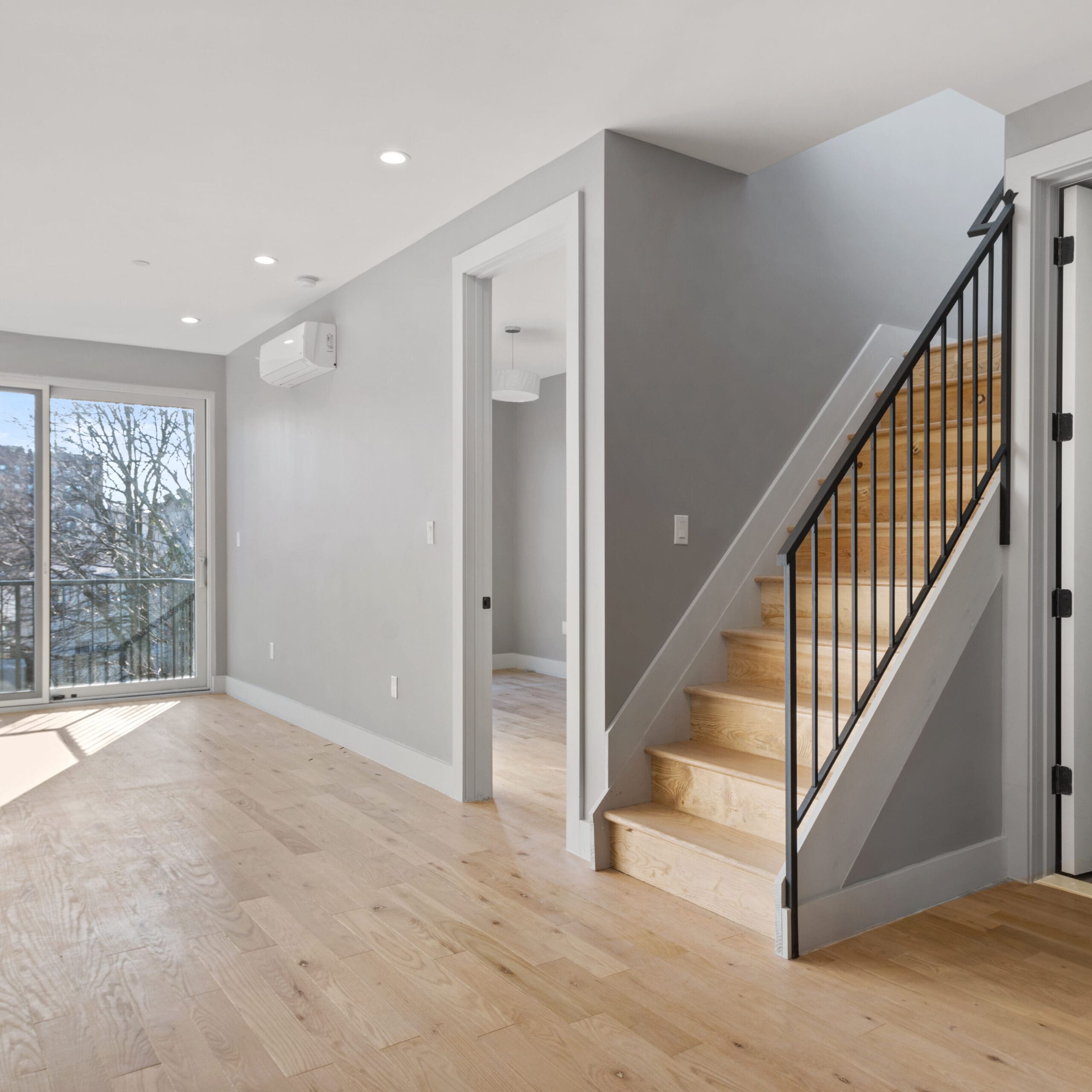 A modern interior space with light-colored hardwood floors, a staircase with wooden steps and a black metal railing, and a room in the background. Large windows to the left let in natural light, illuminating the neutral-toned walls and minimalistic design.