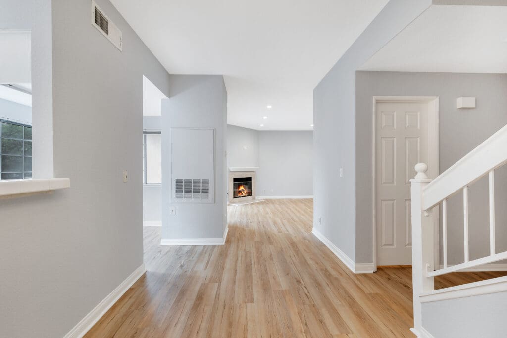 A bright, modern living space with light hardwood flooring and soft gray walls. The room features a small staircase with white railings on the right and a cozy fireplace in the distance. Natural light filters through the windows, creating an airy atmosphere.