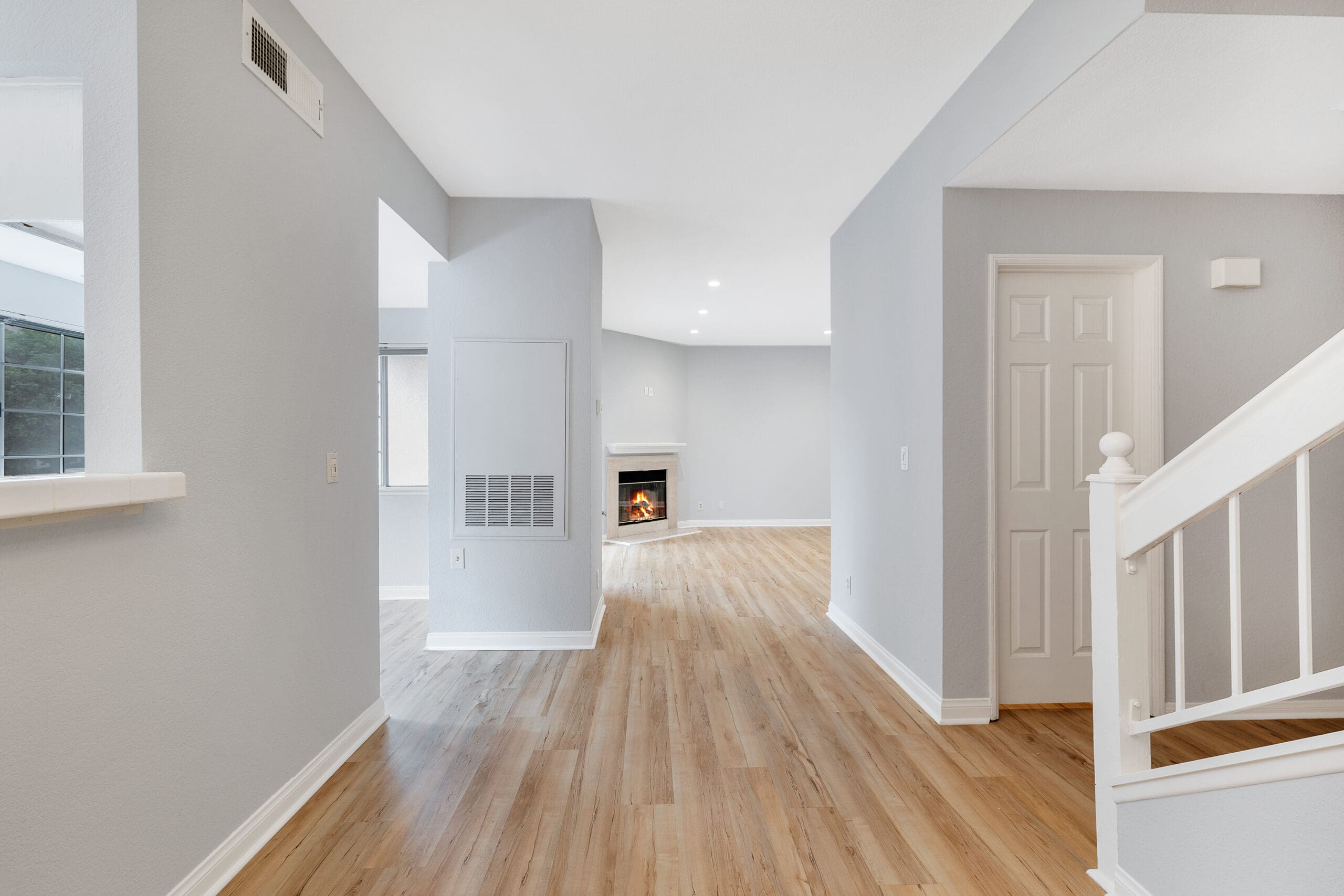 A bright, modern living space with light hardwood flooring and soft gray walls. The room features a small staircase with white railings on the right and a cozy fireplace in the distance. Natural light filters through the windows, creating an airy atmosphere.