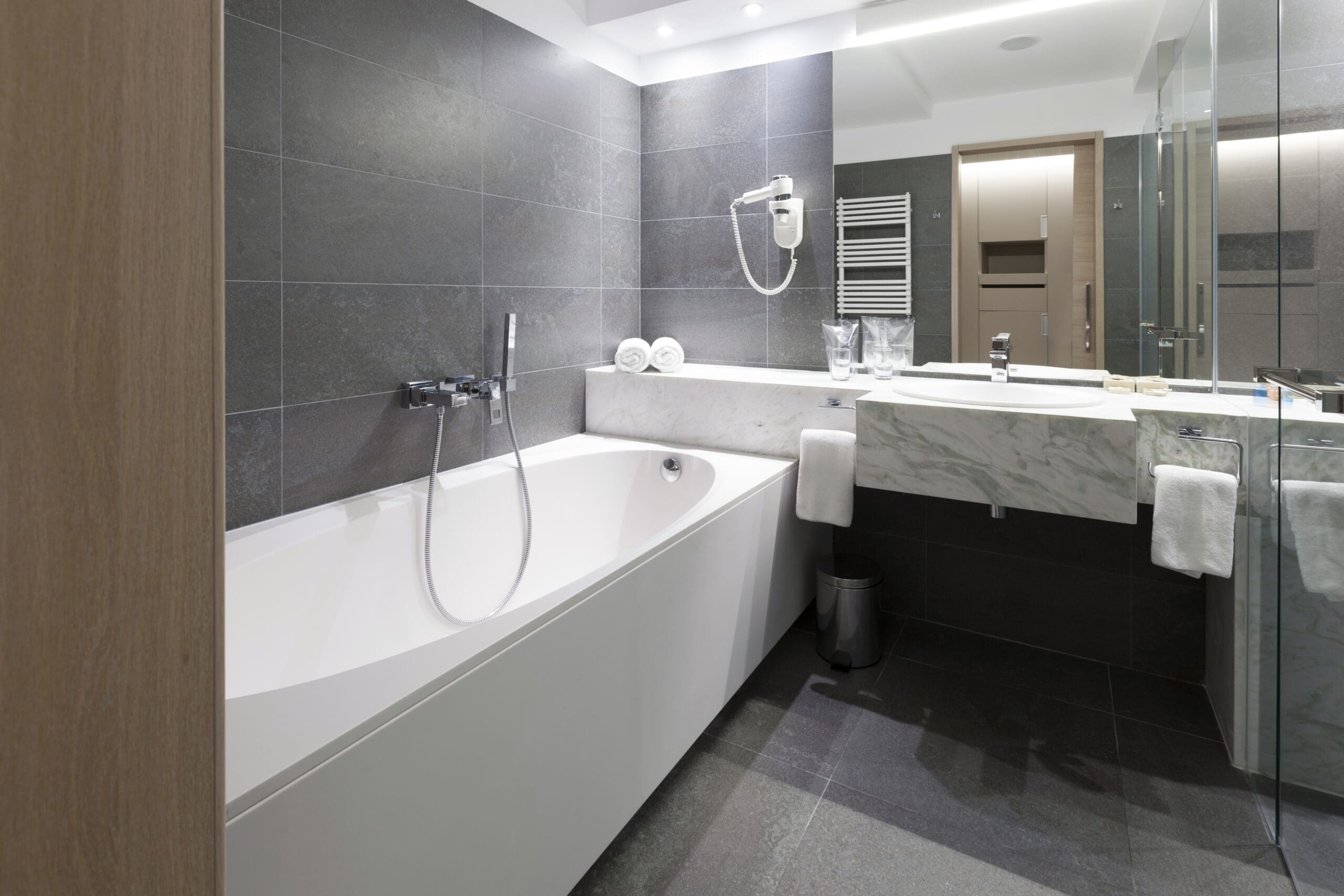 A modern bathroom with gray tile walls and floor, featuring a white bathtub with a handheld showerhead, a marble countertop with a sink, a wall-mounted hairdryer, towel racks, and a glass shower enclosure.