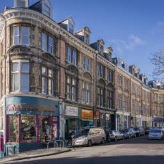 A street corner with a historic multi-story building featuring large windows and intricate architectural details. Shops on the ground floor include a cafe with colorful display windows. Several parked cars line the sloping road, and bare trees are present on the right.