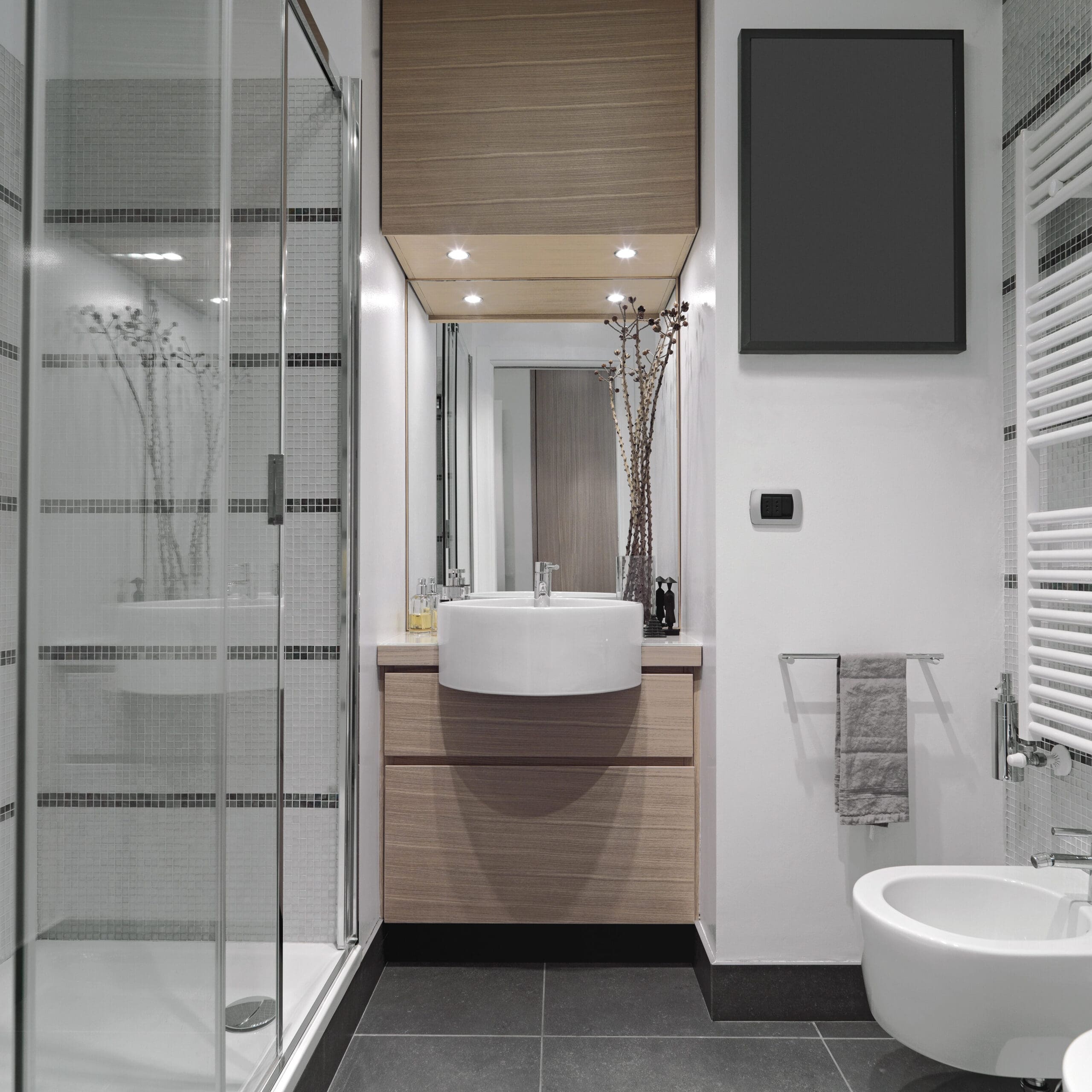 A modern bathroom with a glass-enclosed shower on the left, a wooden vanity with a white vessel sink and a large mirror in the center, and a wall-mounted towel rack with a towel near a white bidet on the right. The bathroom has gray floor tiles and white walls.