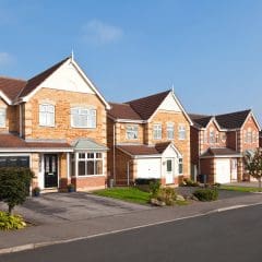 A row of modern, suburban brick houses with peaked roofs and attached garages under a clear blue sky. The houses are neatly maintained, featuring front gardens with green lawns and shrubs. A paved street runs in front of the houses.