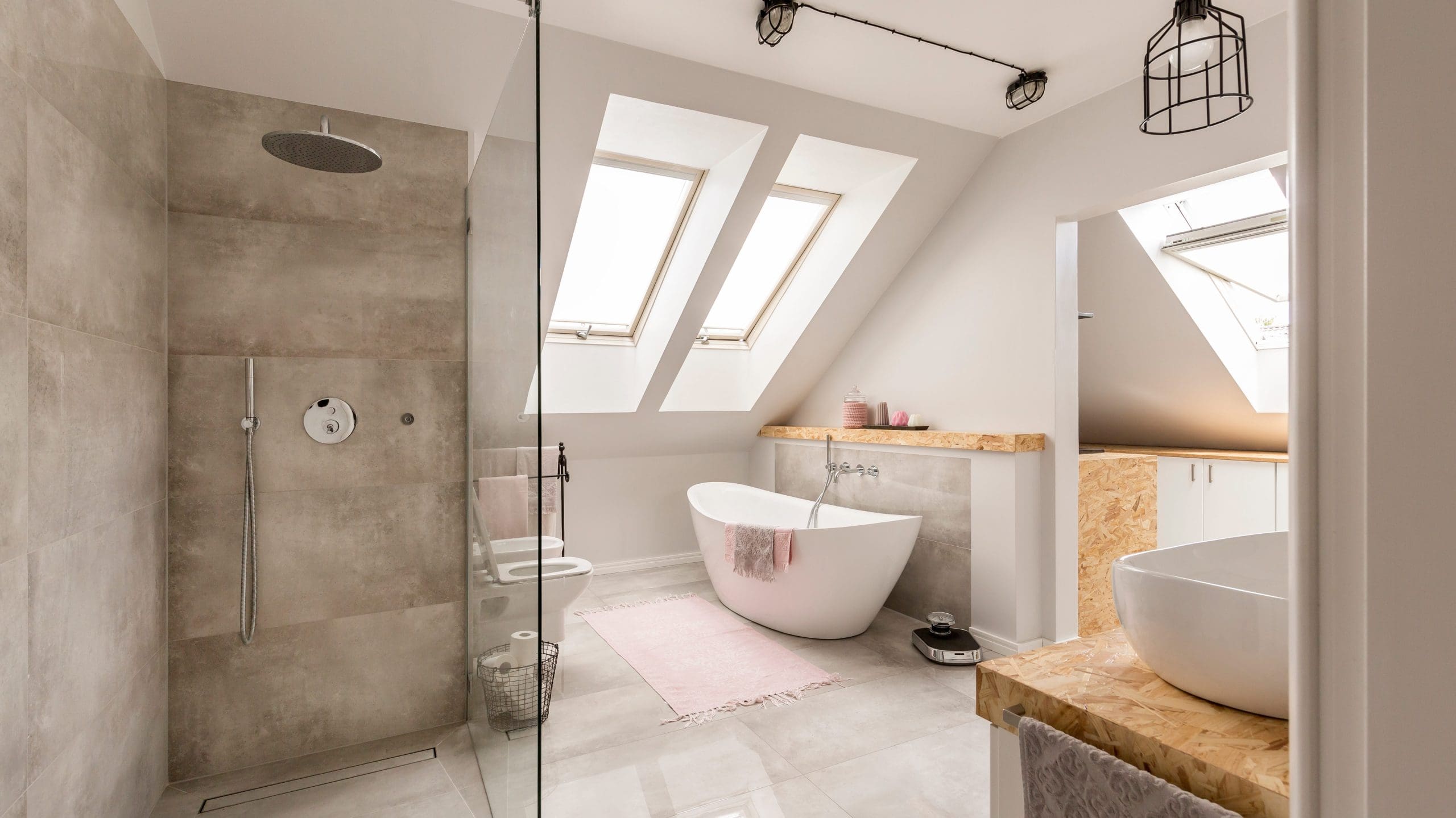 A modern bathroom with large windows, a freestanding bathtub with a pink bath mat, a walk-in shower with a rain showerhead, a white sink on a wooden vanity, and elegant light fixtures. The space is bright and airy with a clean, minimalist design.