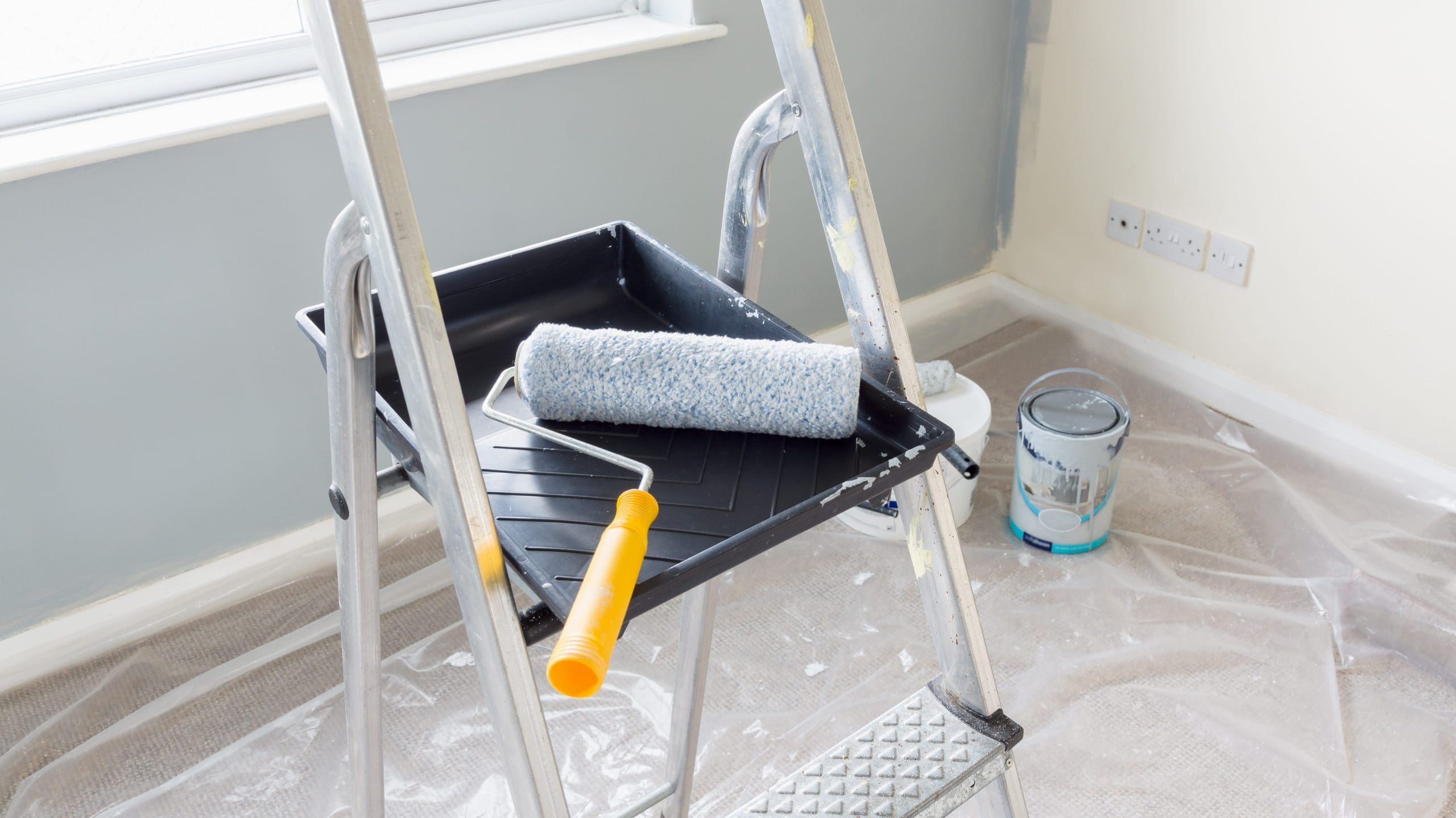 A close-up of a ladder with a paint tray on one of its steps. The paint tray holds a paint roller with a yellow handle covered in gray paint. Nearby, there are paint cans on a plastic-covered floor in a room with partially painted gray walls.