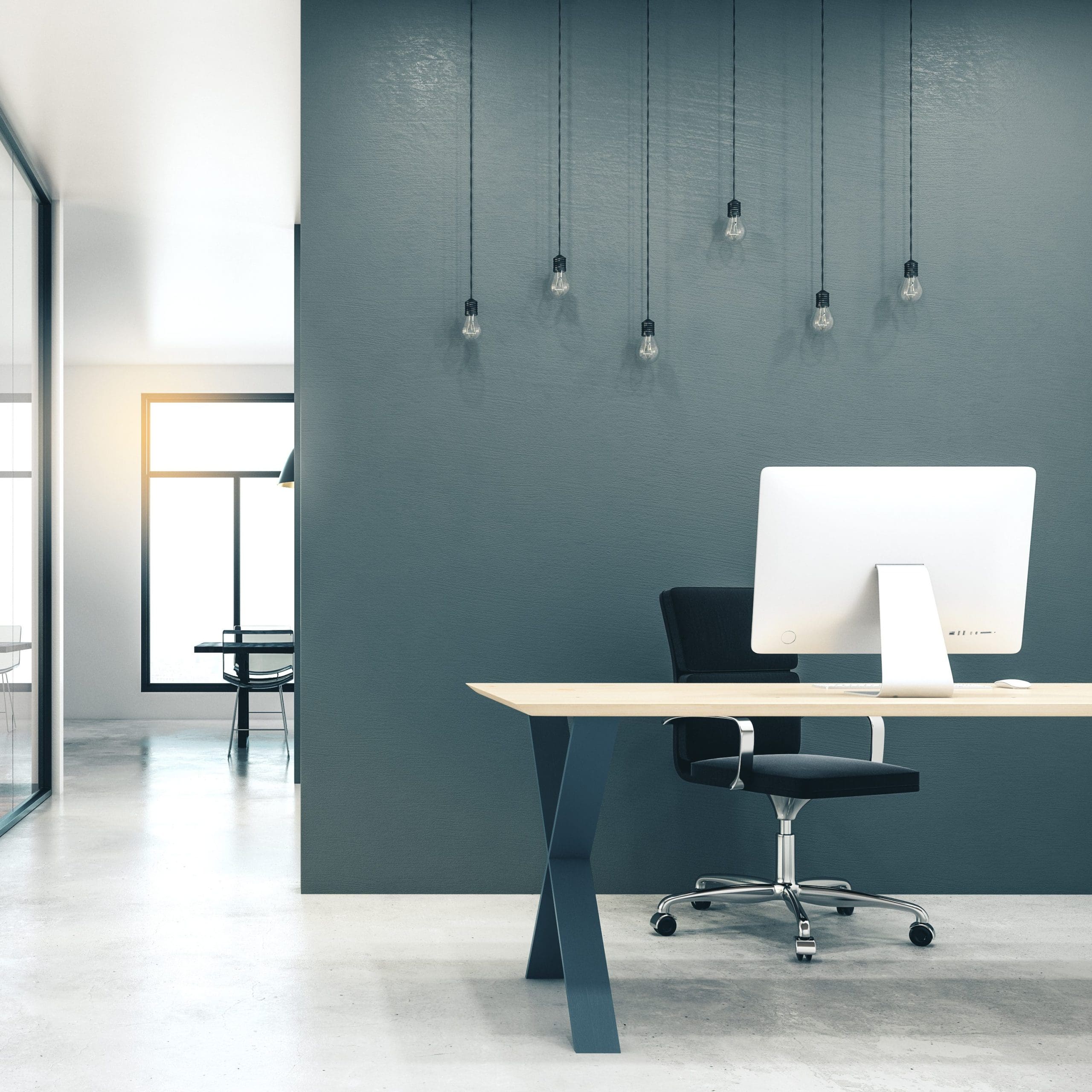 Minimalist office space with a clean, modern design. Features a white desktop computer on a light wood desk, a black office chair, and a gray wall adorned with hanging exposed light bulbs. Large windows in the background allow natural light to brighten the room.