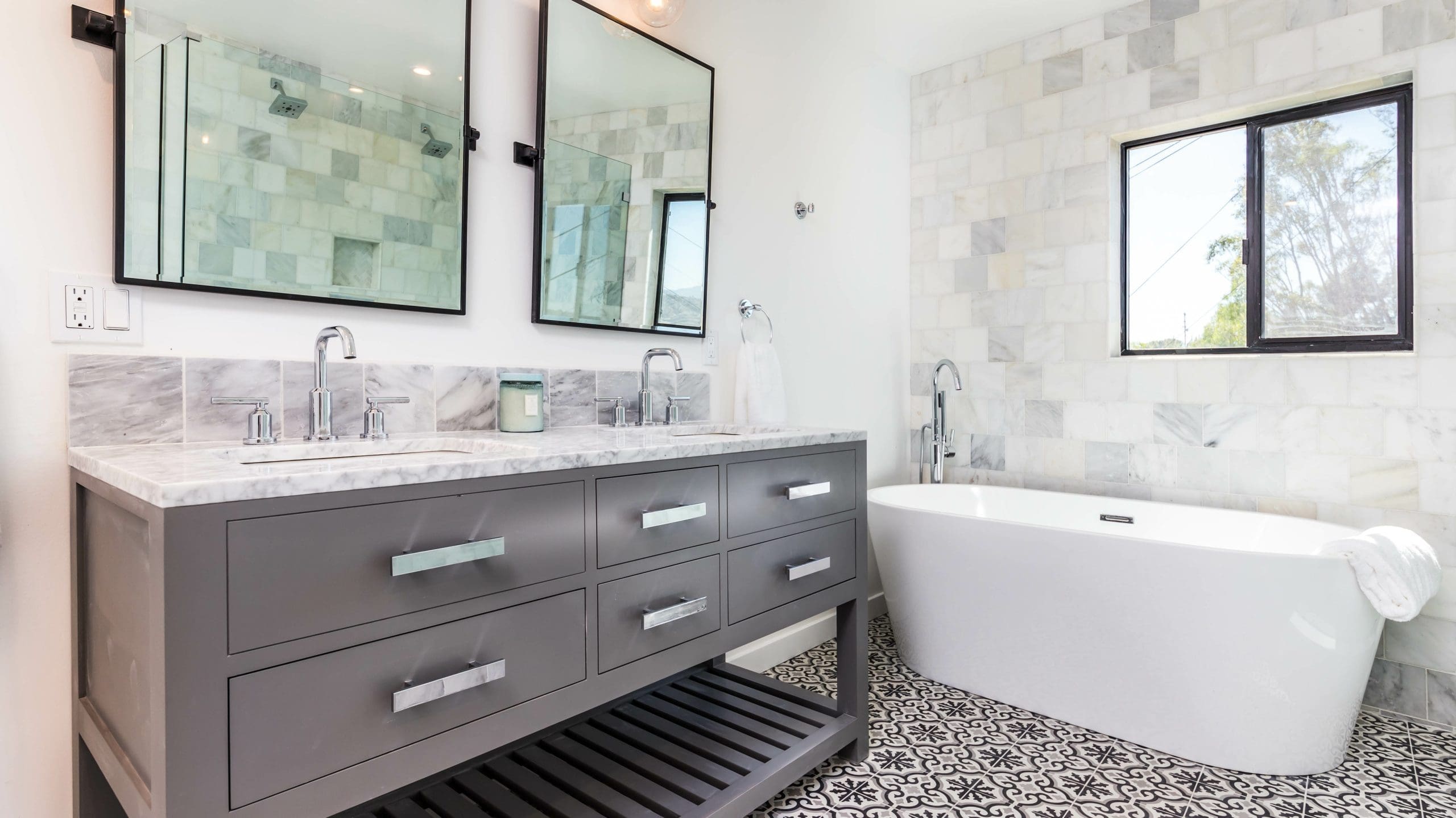 A modern bathroom with a marble-topped double vanity featuring two mirrors and silver faucets. A freestanding white bathtub is positioned by a window with patterned floor tiles and white tiled walls. A towel is draped over the bathtub edge.