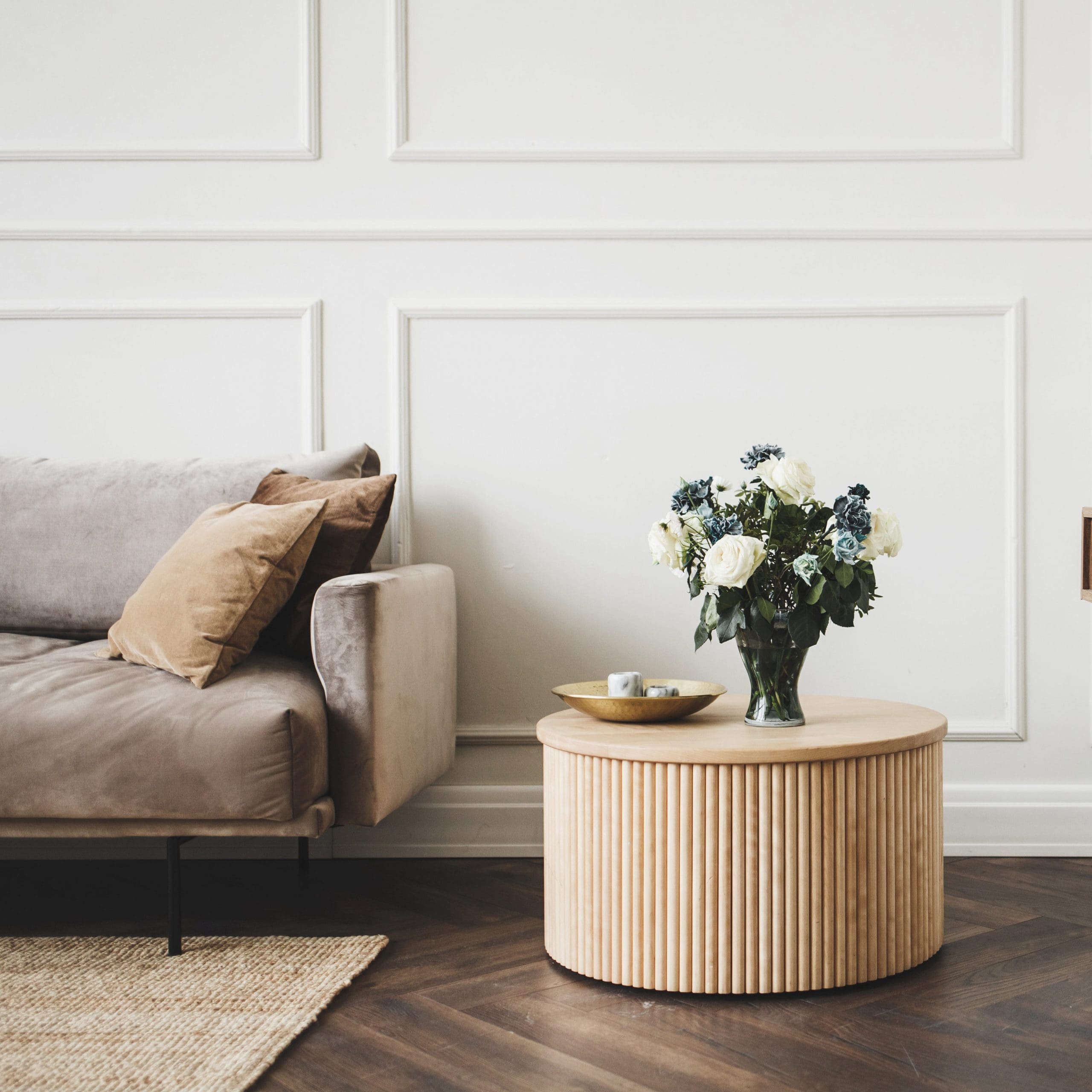 A minimalist living room with a gray sofa adorned with two beige cushions, next to a round wooden coffee table. The coffee table displays a bouquet of white and blue flowers in a vase and a decorative tray with items. The room has a herringbone wooden floor and paneled walls.