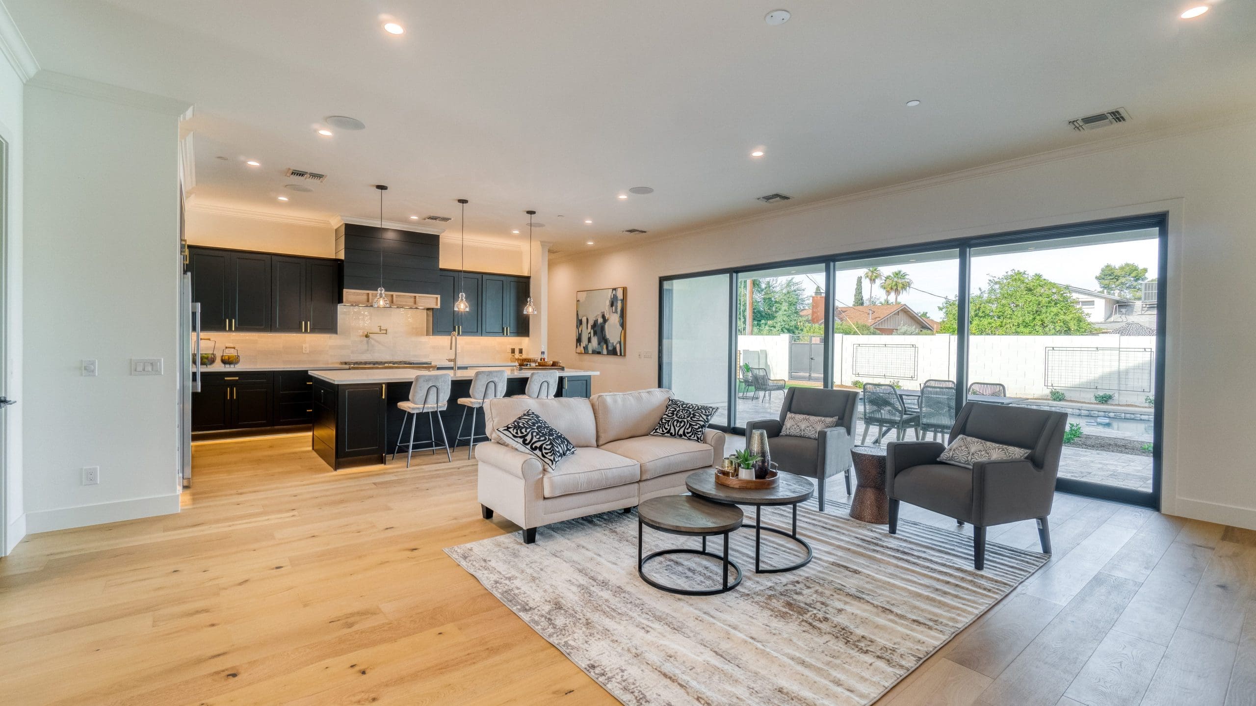 A modern open-concept living room and kitchen with light wood flooring. The living room has a light-colored sofa, two dark armchairs, and a coffee table on a patterned rug. The kitchen features dark cabinets, a large island with barstools, and stainless steel appliances. Sliding glass doors lead to an outdoor patio.