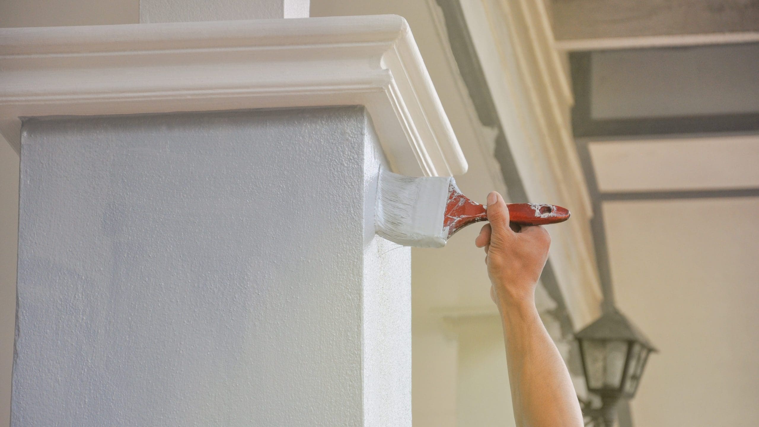 A person's hand holding a paintbrush applies white paint to a gray column. The column features a square shape with detailed molding near the top. A lantern-style light fixture is partially visible in the background.