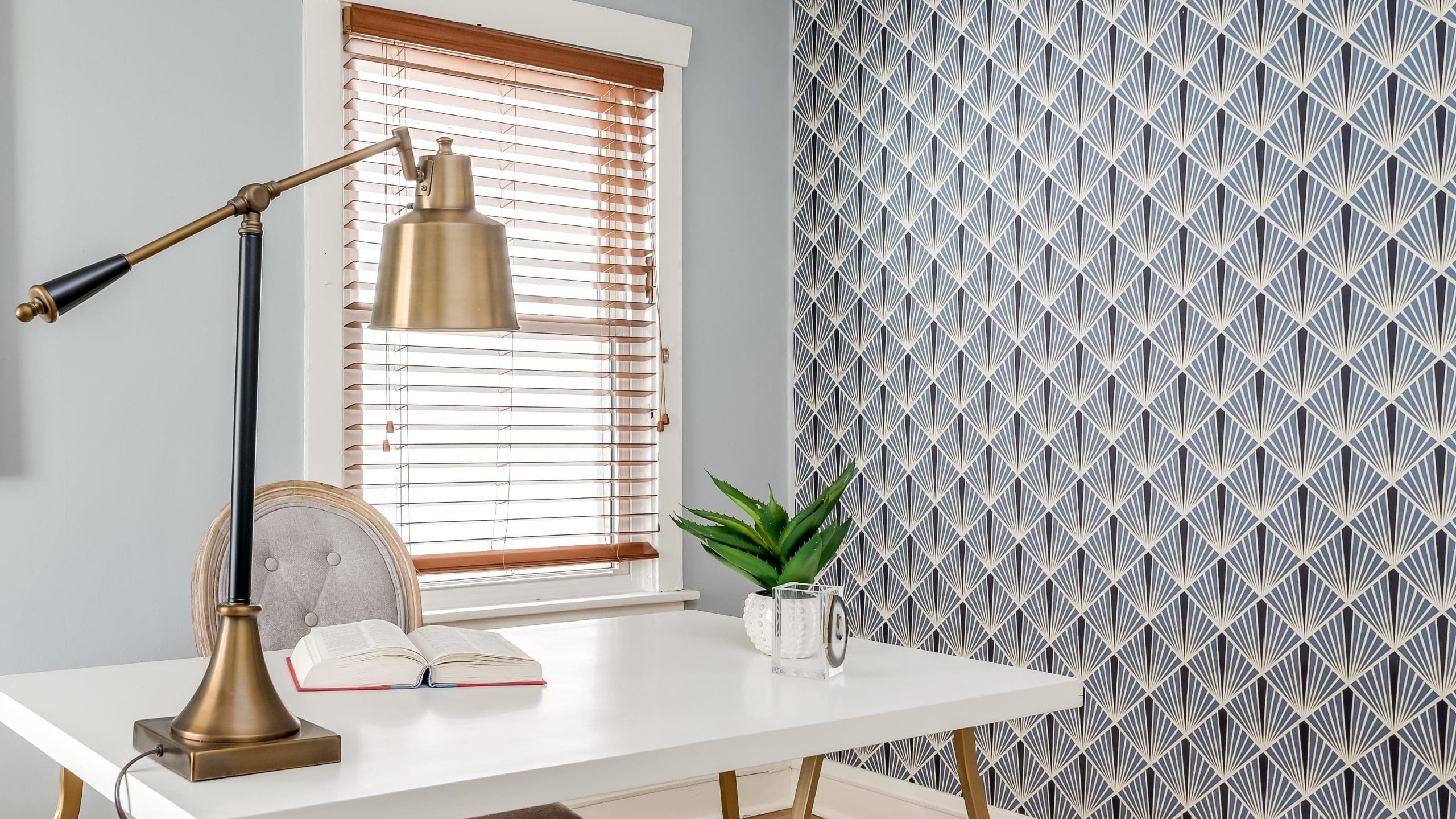 A modern home office with a white desk featuring an open book, a brass desk lamp, and a potted plant. Light blue wallpaper with a geometric diamond pattern adorns one wall, while a window with wooden blinds provides natural light. Pale blue-gray walls complete the decor.