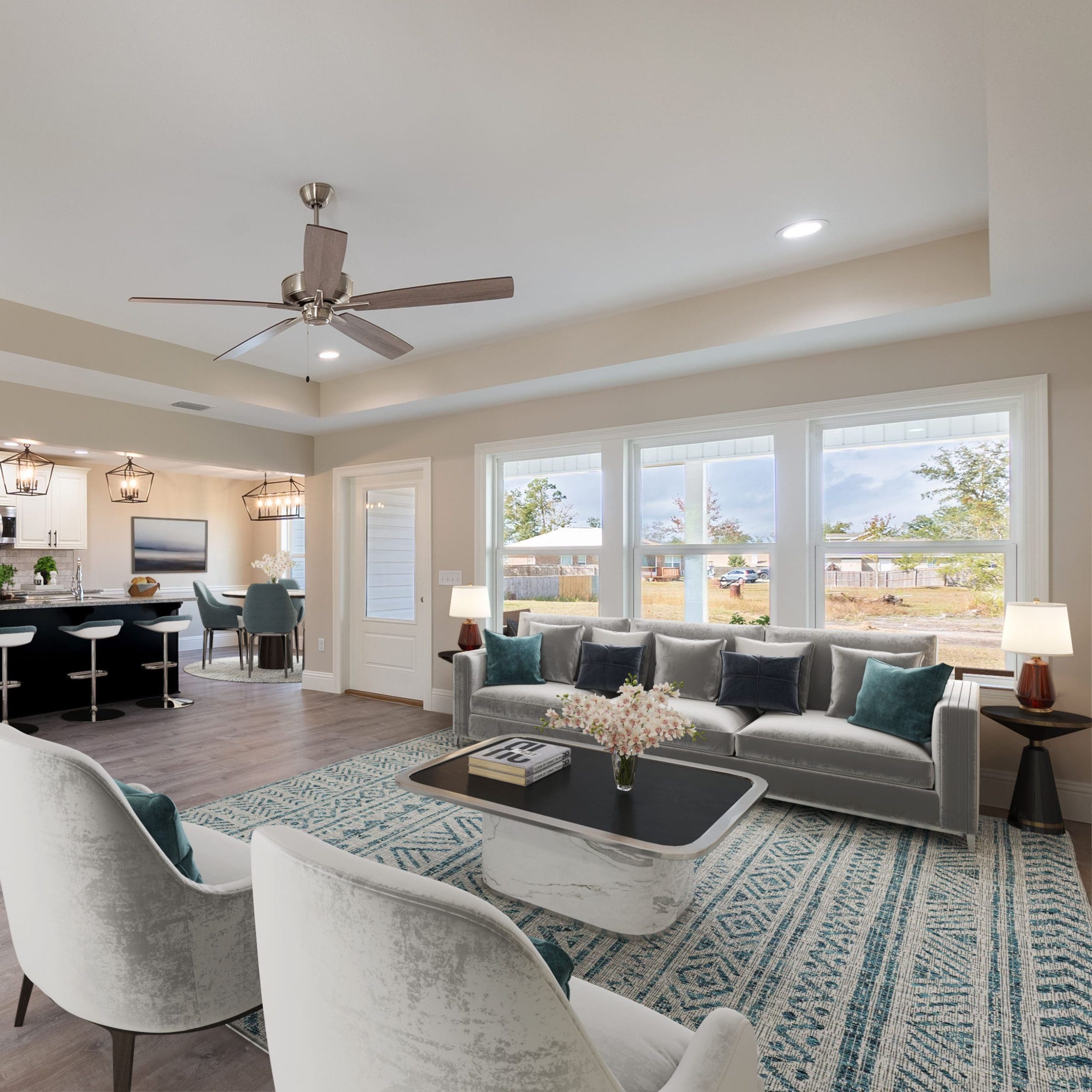 A bright, modern living room with large windows letting in natural light. The space is furnished with a gray sofa, two white armchairs, a patterned area rug, and a coffee table. The open layout extends to a kitchen with a breakfast bar and stools in the background.