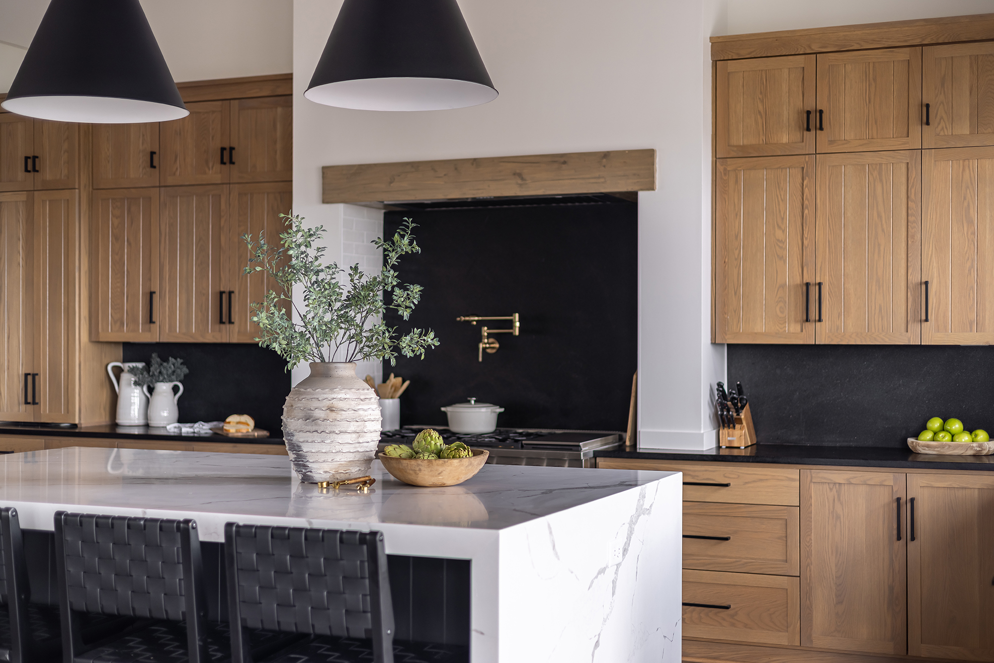 A modern kitchen featuring light wood cabinets, black countertops, and a central island with a white marble surface. A vase with greenery and a bowl of fruit adorn the island. Black chairs line the island, and pendant lights hang above.