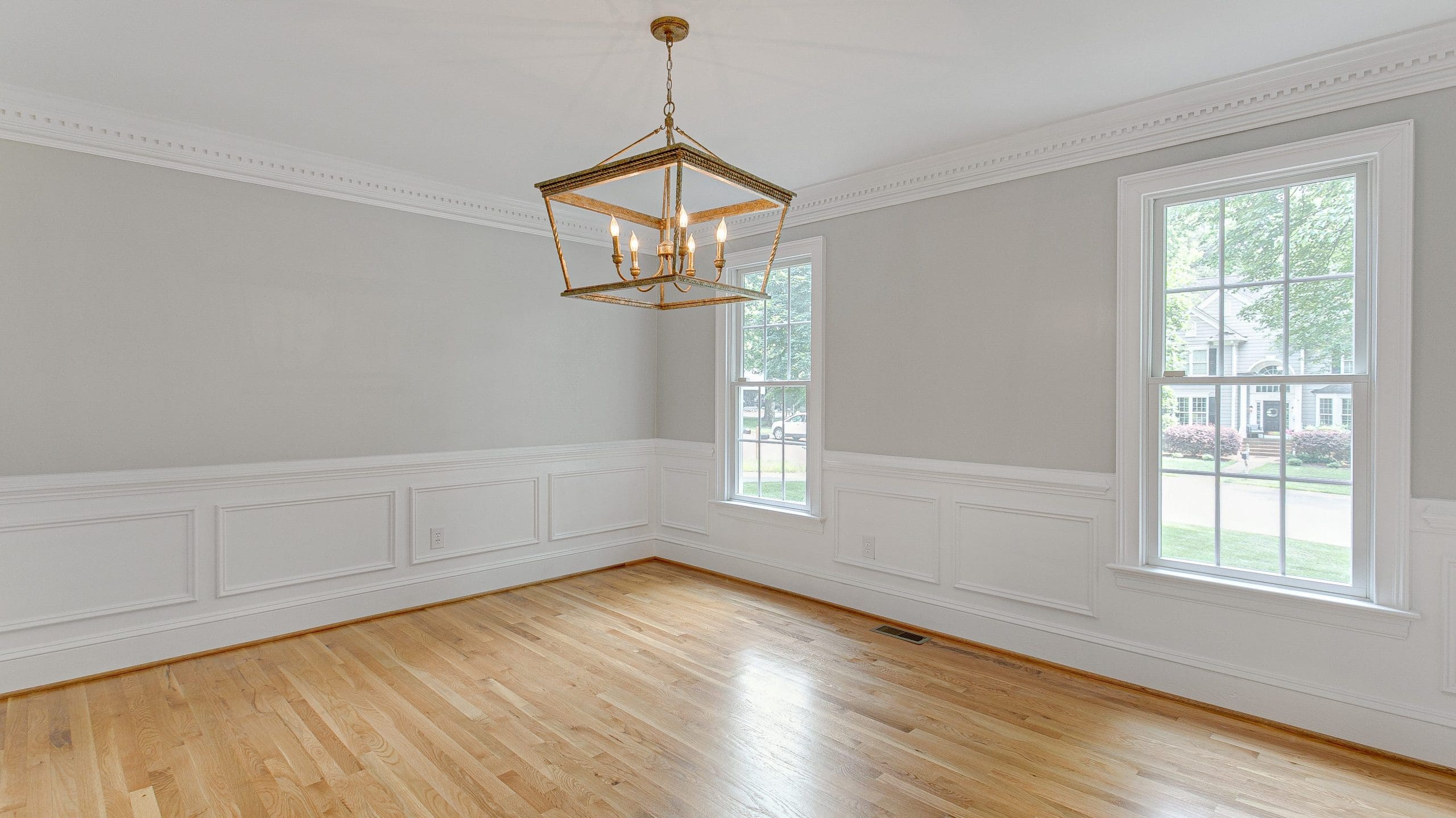 A bright, empty room with light gray walls, white wainscoting, and a polished hardwood floor. Two large windows allow natural light in, and a modern gold and glass chandelier hangs from the ceiling. The windows offer a view of a tree-lined street outside.