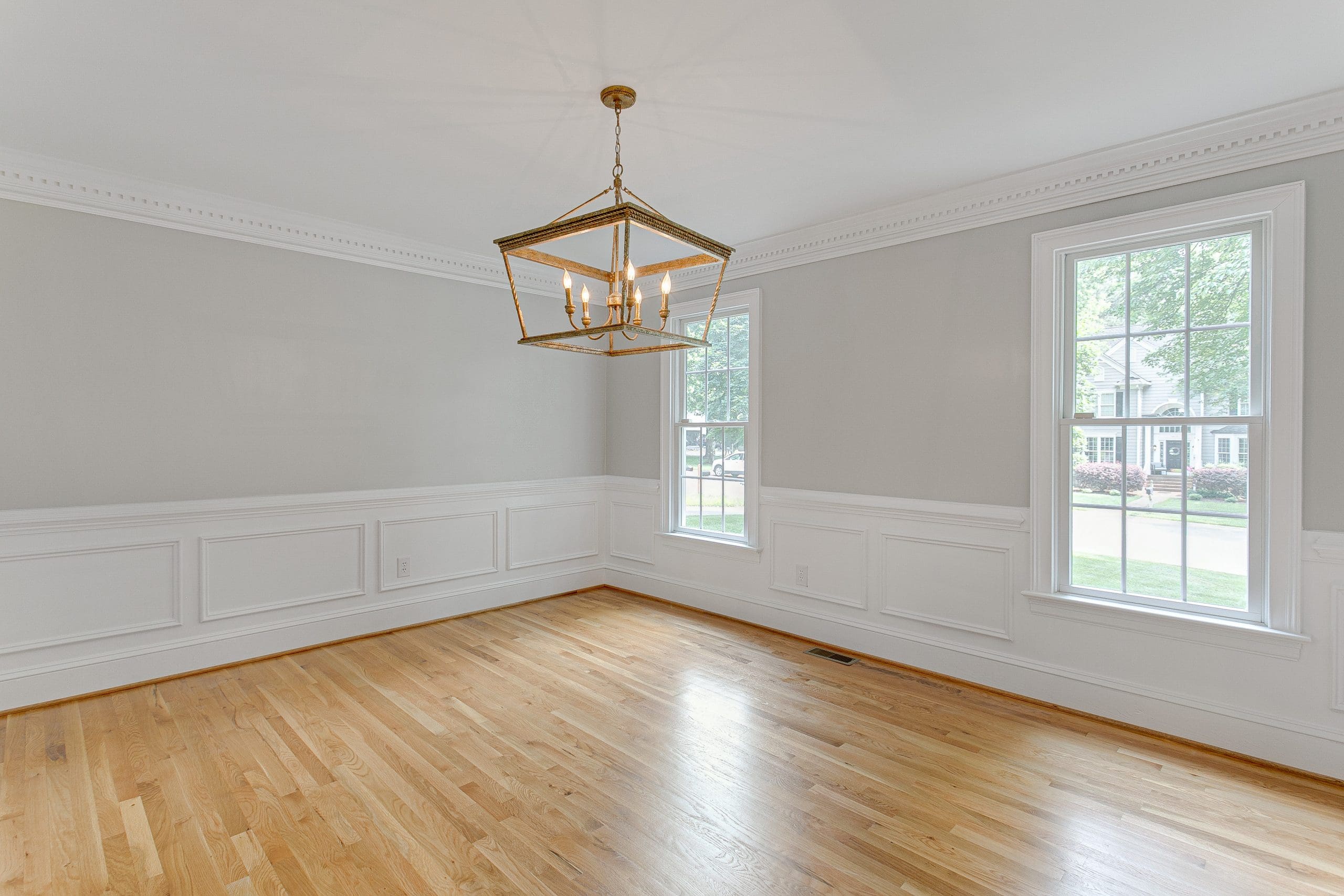 A vacant room with light gray walls, white wainscoting, and wooden flooring. Two large windows on the right wall let in natural light. A gold-colored, geometric chandelier hangs from the ceiling in the center of the room. Trees and houses are visible through the windows.