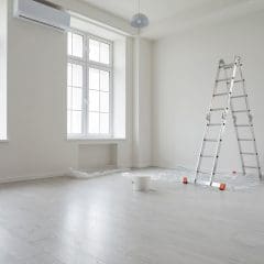 A bright, empty room with light wooden flooring. Large windows let in natural light. A folded aluminum ladder stands in the corner next to a paint tray and a plastic sheet. A white paint bucket sits on the floor, indicating ongoing painting work.