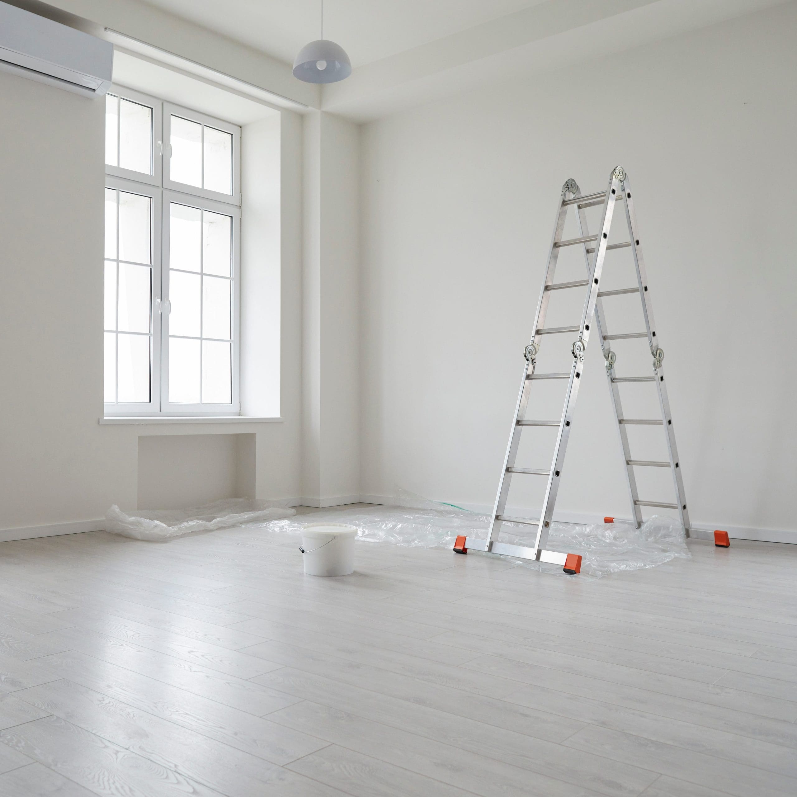 A minimal, empty room with light wooden flooring, a large window, and white walls. A metal ladder stands on a plastic sheet spread across the floor, next to a small white paint bucket, indicating a room in the process of being painted or renovated.