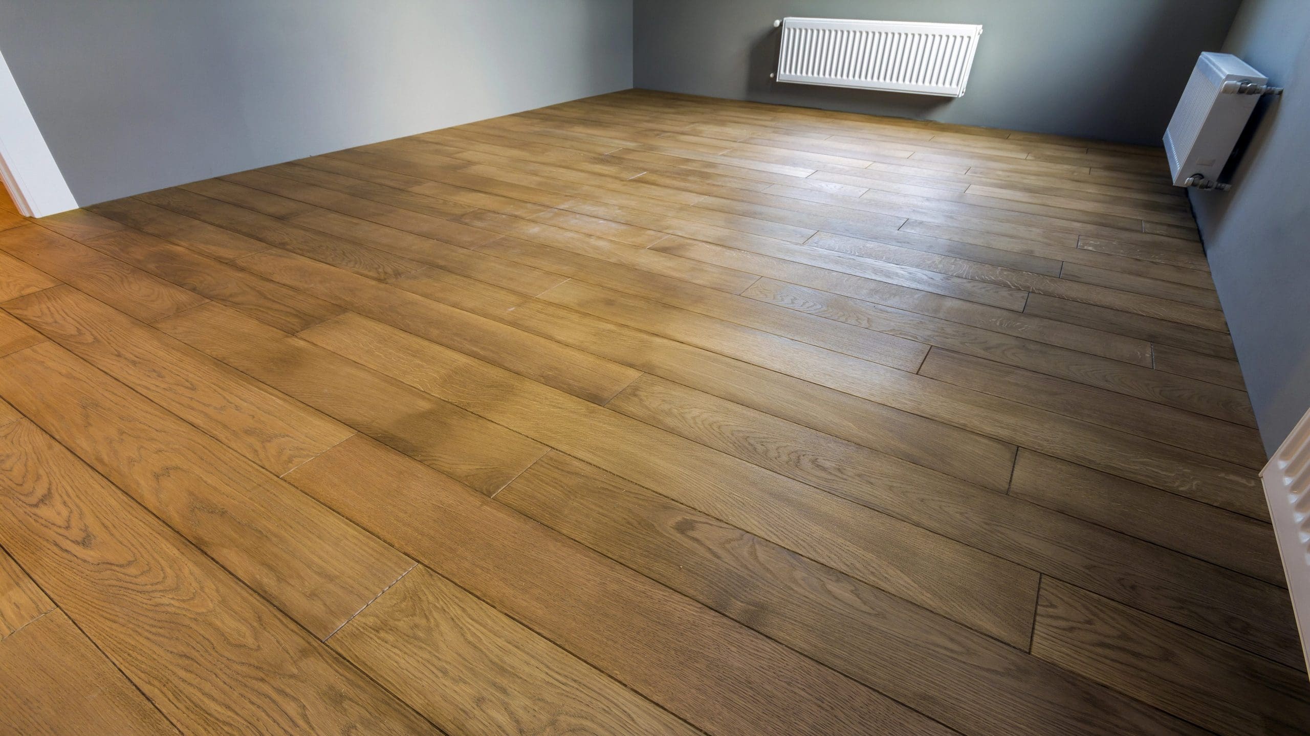 A spacious, empty room with wooden flooring and gray walls. A white radiator is attached to one of the walls, and there is no furniture or decoration in the room. The wooden floorboards have a natural finish and are arranged in parallel rows.