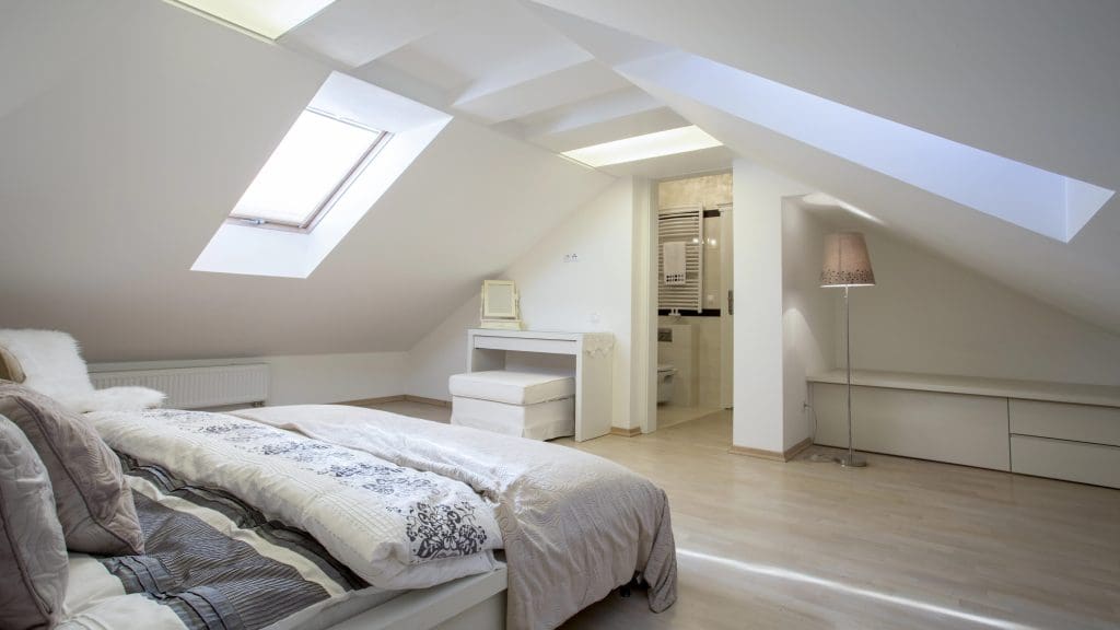 A modern attic bedroom with sloped ceilings featuring large skylights. The room includes a neatly made bed with white and patterned bedding, a small dressing table with a stool, minimalistic decor, and an en-suite bathroom with a visible sink area.