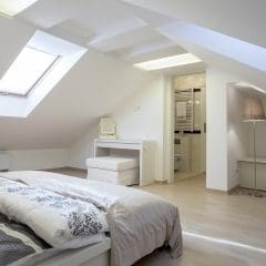 A modern attic bedroom with sloped ceilings featuring large skylights. The room includes a neatly made bed with white and patterned bedding, a small dressing table with a stool, minimalistic decor, and an en-suite bathroom with a visible sink area.