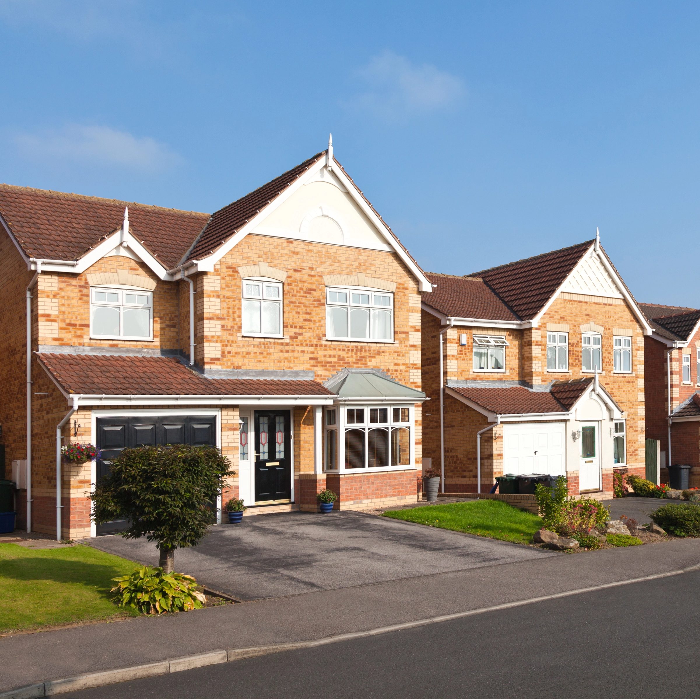 A suburban street with two detached, modern brick houses. Each house has a garage, a well-maintained front garden, and large windows. The sky is clear and blue, indicating sunny weather.