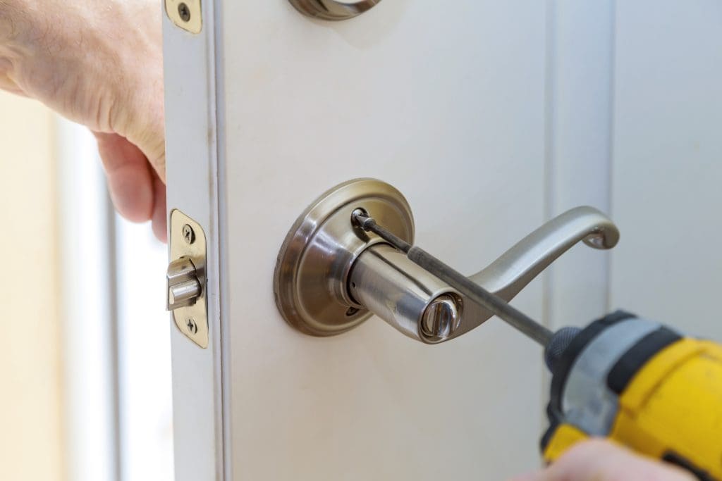 A person using a power drill to install a metallic door handle on a white door. The hand is holding the door steady while tightening a screw on the handle. The door latch and lock mechanism are visible.
