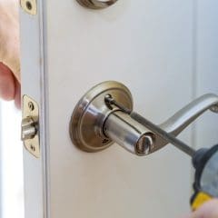 A person using a power drill to install a metallic door handle on a white door. The hand is holding the door steady while tightening a screw on the handle. The door latch and lock mechanism are visible.