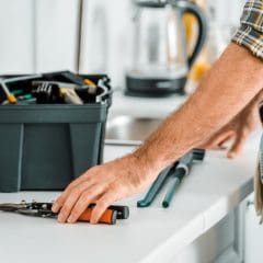 A person in a plaid shirt reaches for a pair of pliers on a white counter, with a toolbox filled with tools and a drill nearby. The individual's tool belt is visible, containing various hand tools. A kettle and sink are seen in the background.