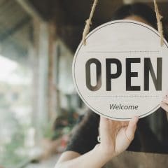 A person is hanging a circular sign that says "OPEN Welcome" on a glass window. The background shows a blurred interior of what appears to be a store or café. The view outside the window is also slightly visible, showing greenery and buildings.