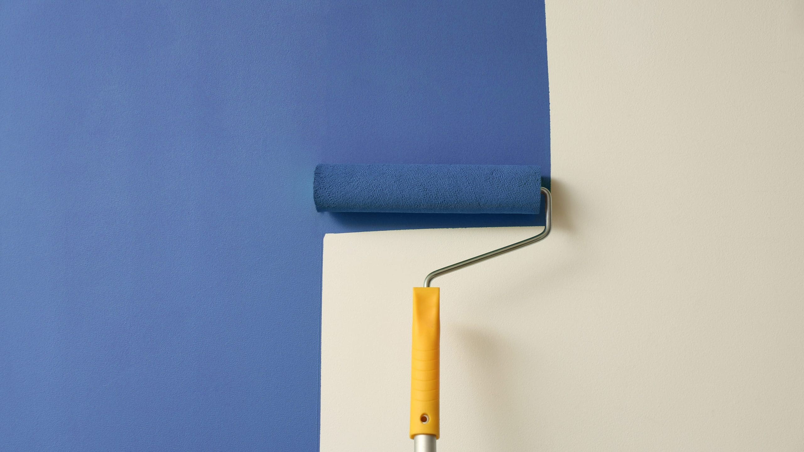 A paint roller with a yellow handle is being used to apply blue paint on a light, nearly white wall. The painting process is halfway completed, with a clear distinction between the painted and unpainted sections.