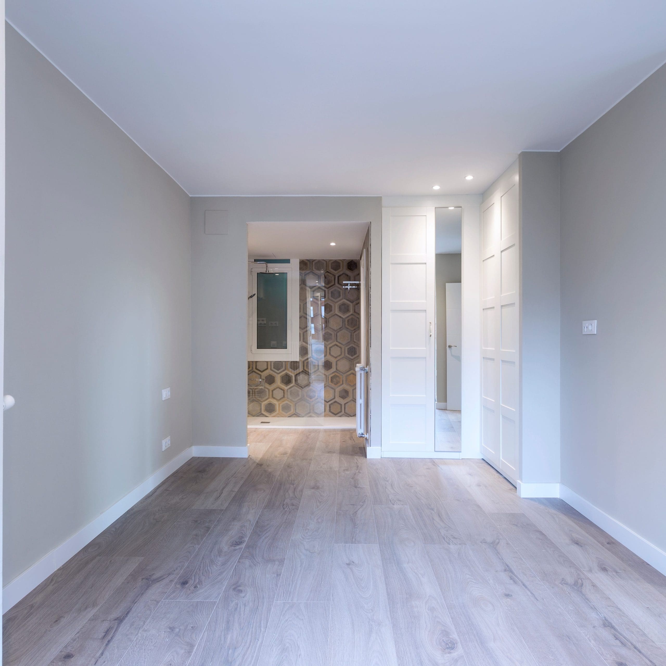 A bright, empty room with light wood flooring, gray walls, and recessed lighting. To the back is an open doorway leading to a small area with hexagonal patterned tiles and a glass shower door. White double doors and built-in closets are visible on the right.