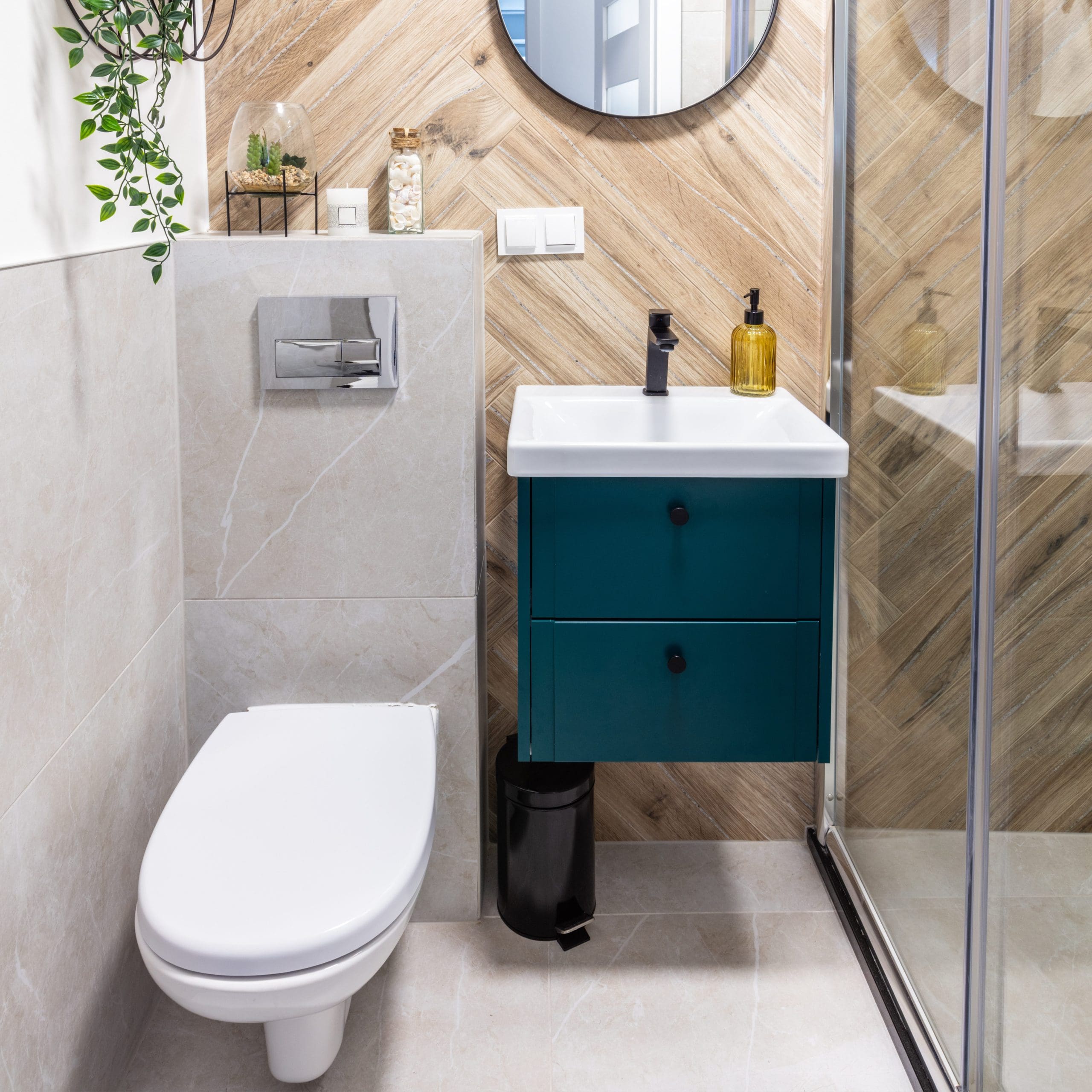 A modern bathroom with a wall-mounted toilet, a small sink with a teal cabinet underneath, and a sleek black faucet. The shower enclosure has a glass door, and the walls feature light grey tiles with a wooden accent wall. A circular mirror hangs above the sink.