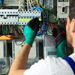 A technician wearing green gloves and a white shirt works on an electrical panel filled with various wires and circuit breakers. He is using tools to adjust connections, focusing closely on his task.