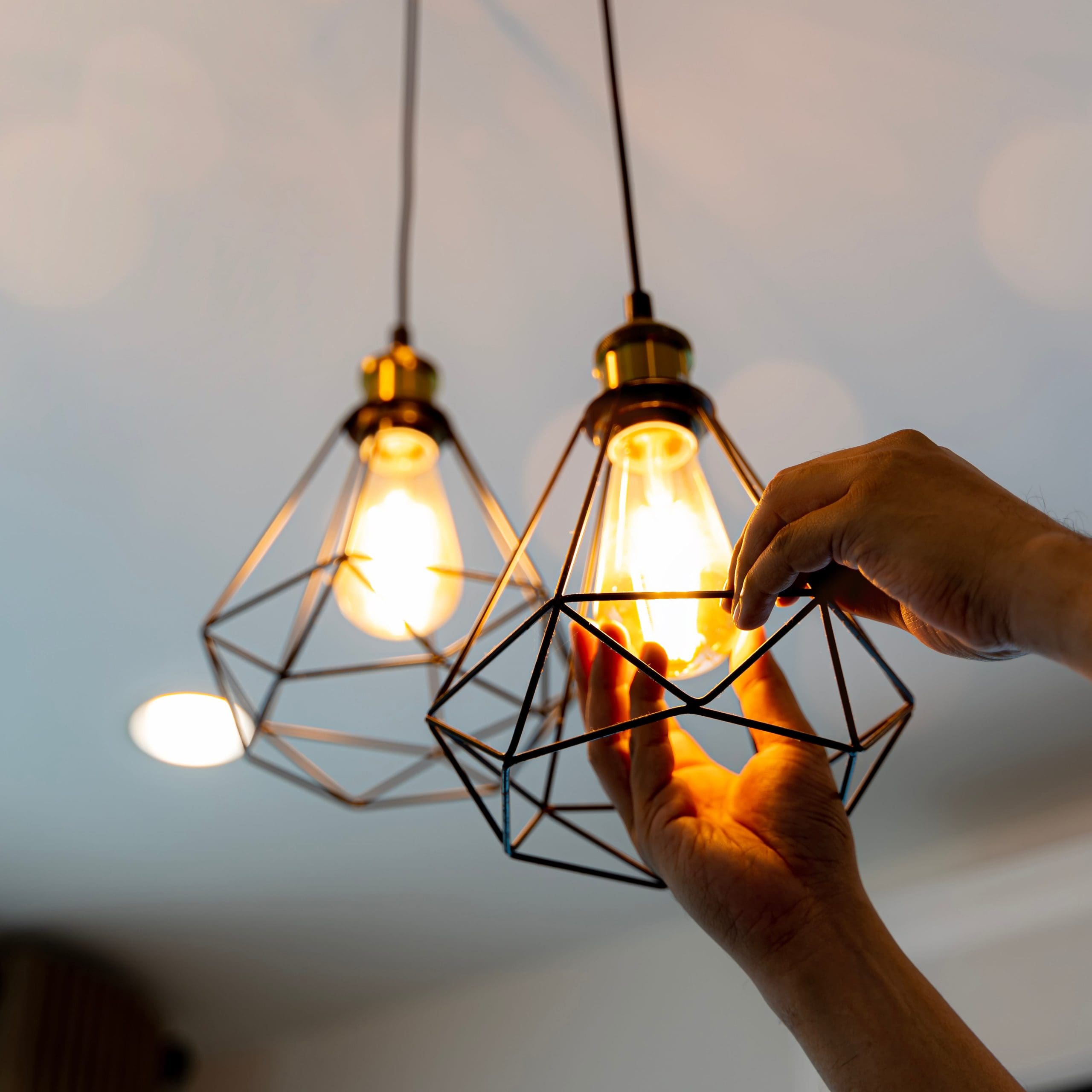 Two modern pendant light fixtures with geometric wire frames hang from the ceiling. One light bulb is illuminated, while a person's hands are adjusting the bulb in the other fixture, causing it to emit a soft glow. The background is softly lit.