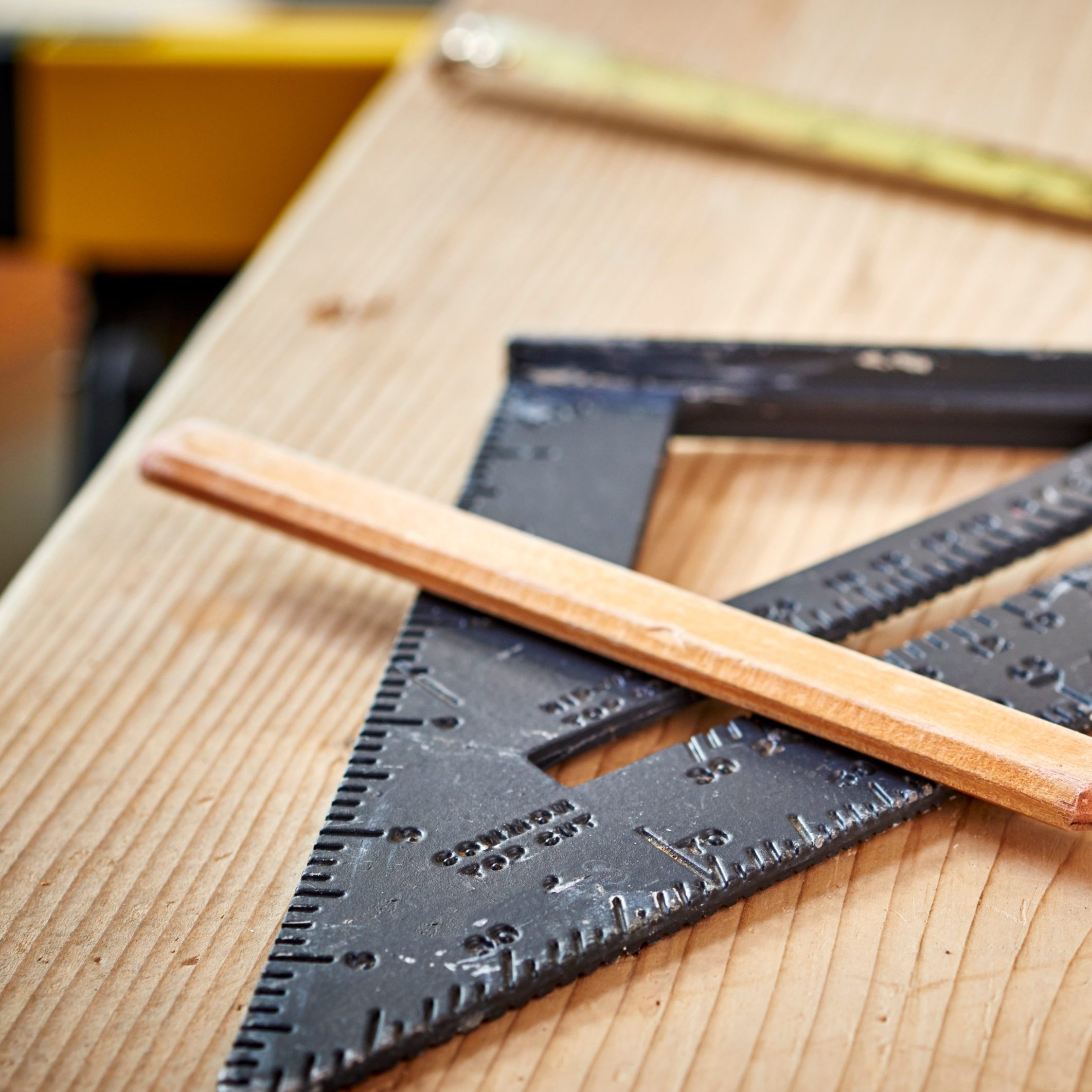 A close-up image of a drafting triangle and a wooden pencil lying on a wooden table. The drafting triangle is black with ruler markings, and part of a yellow tape measure can be seen in the background. The table has a light natural wood grain texture.