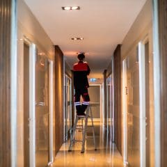 A person in high-visibility clothing stands on a ladder fixing a ceiling light in a brightly lit corridor. The corridor has polished floors and wood-paneled walls, with doors on both sides leading to rooms. Ceiling lights illuminate the hallway.