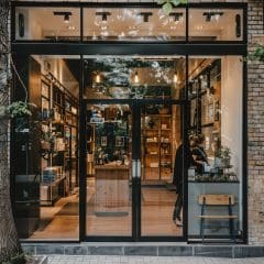 A modern storefront with large glass windows and a door, revealing a cozy and well-lit interior featuring wooden shelves and displays. A person inside is browsing items. Outside, there are potted plants and a tree in the foreground.