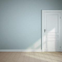 An empty room with light blue walls, white baseboards, and a closed white door. Sunlight streams in from an unseen window, casting a bright beam across the door and the light wood chevron-patterned floor. The room has a calm and minimalist ambiance.