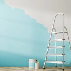 A step ladder stands in front of a wall partially painted in a light blue color. Two paint cans and a paintbrush rest on the floor nearby. The lower part of the wall is painted blue, while the upper section remains white. The floor has a wood pattern.