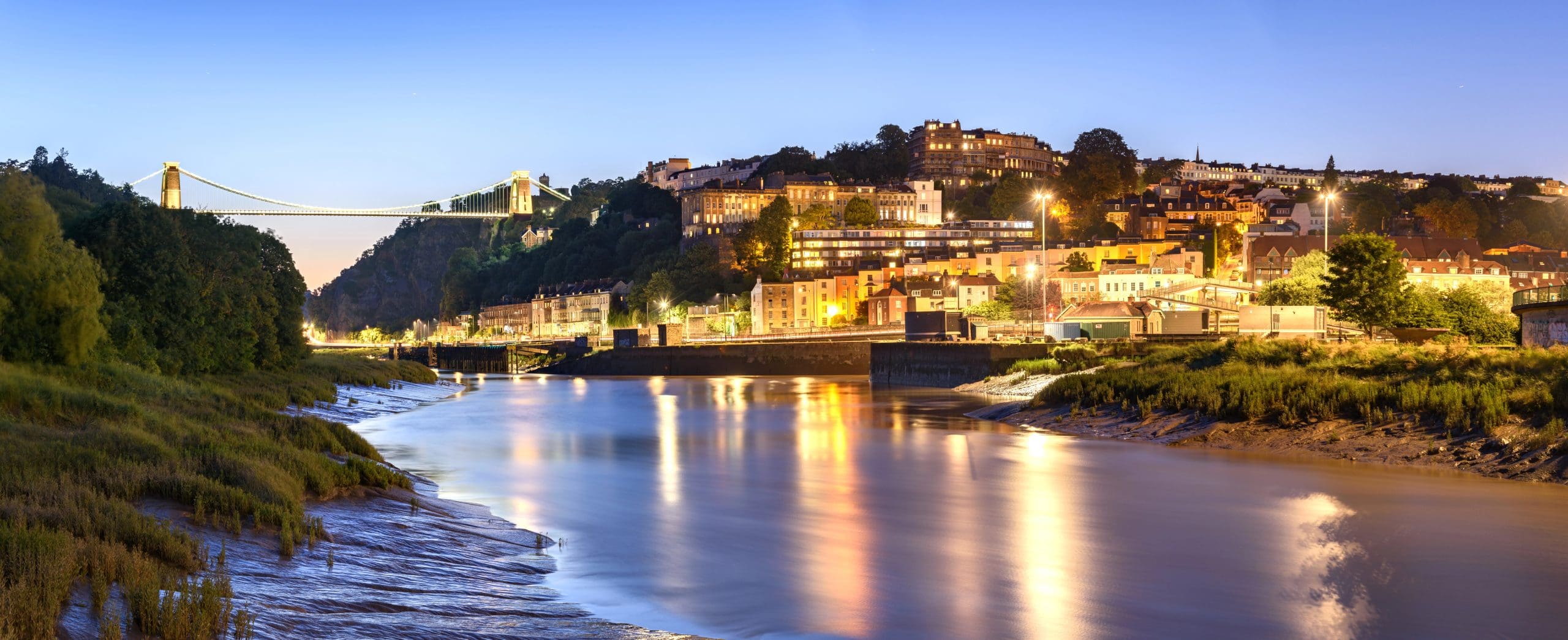 A panoramic view of a cityscape at dusk featuring a river with reflections of lights from buildings along the shore. A suspension bridge is seen on the left spanning across the river, with illuminated buildings and greenery on the hillside in the background.