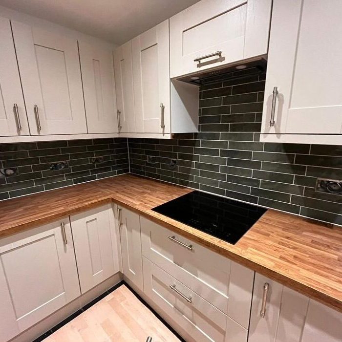 A modern kitchen corner with white cabinets and drawers, wooden countertops, and a black electric stovetop. The backsplash features dark green, glossy subway tiles. The floor is a light wood tone, and there are stainless steel handles on the cabinetry.