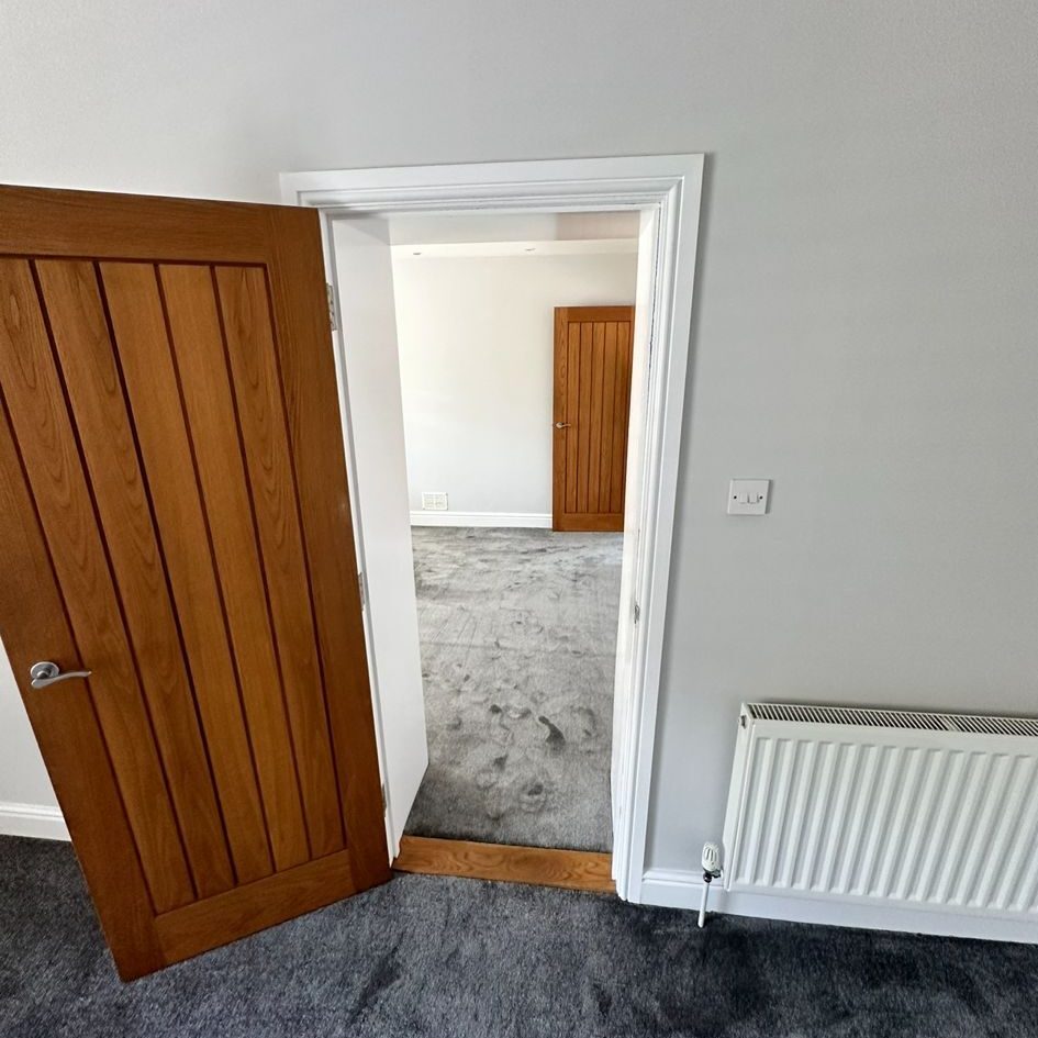 A room with grey carpet has a wooden door that is open, leading to another room with a grey carpet. The walls are white, and a white radiator is mounted on the wall next to the door. The far room also has a closed wooden door.