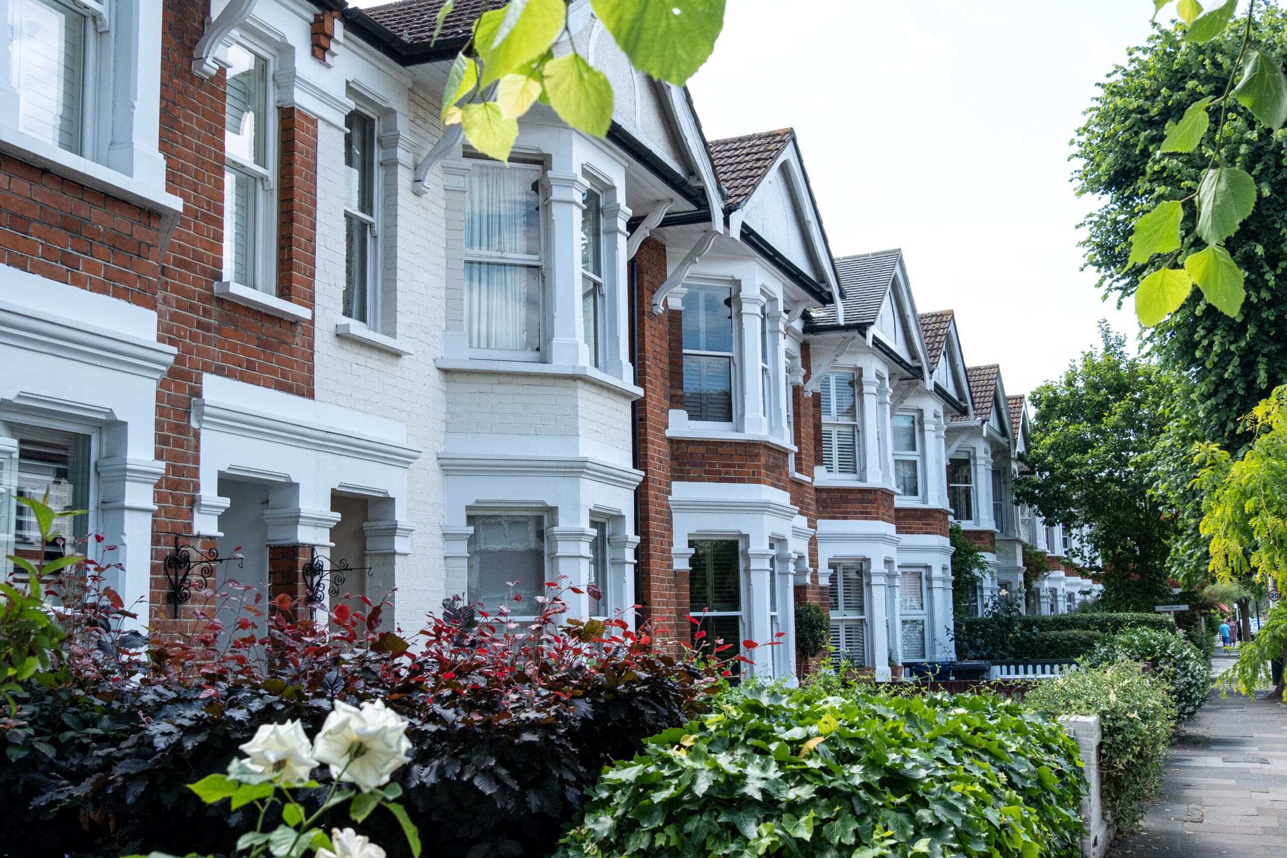 A row of traditional British terraced houses with red brick and white trim, surrounded by green bushes and trees, on a quiet residential street.
