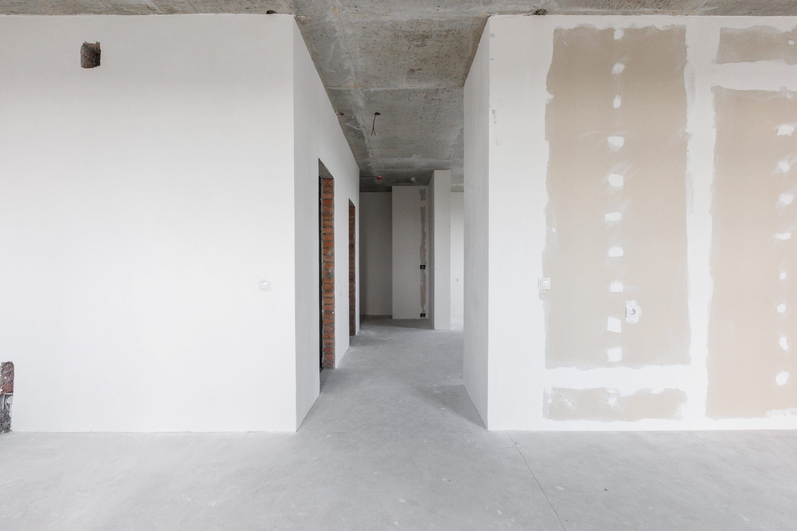 A partially finished interior with white walls, exposed concrete ceiling, and unfinished drywall. Doorways lead to other rooms, and construction materials are visible, indicating ongoing renovation or construction work.