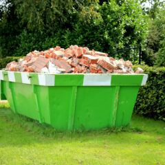 A large green dumpster filled with broken red and white bricks sits on a grassy lawn, surrounded by bushes and trees on a sunny day.