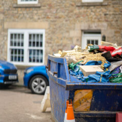 A large blue dumpster filled with construction debris is positioned in front of a stone building. Two orange traffic cones stand nearby, and blue cars are parked along the street.