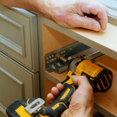A person uses a yellow cordless drill to install a metal drawer slide inside a wooden cabinet. Another person holds the edge of the cabinet steady.